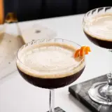 Due cocktail scuri con schiuma chiara, guarniti con scorza d'arancia. Un'idea per un caffè e succo d'arancia rivisitato.
