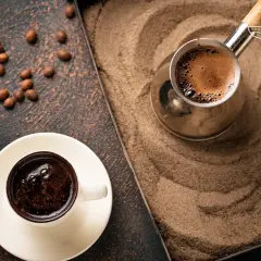 Un caffè arabo fumante in una tazzina bianca, accanto a un bricco di metallo su sabbia calda. Chicchi di caffè sparsi.