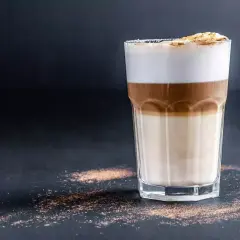 Un bicchiere di caffè macchiato caldo con strati di latte e caffè, sormontato da schiuma soffice e una spolverata di cacao.