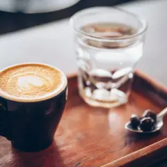 Tazza di caffè con latte art e bicchiere d'acqua, accompagnati da chicchi di caffè su un cucchiaino, su un vassoio di legno.