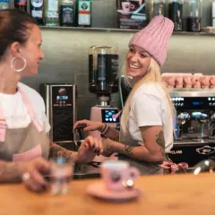 Due donne sorridono dietro il bancone di un bar, una con un cappello rosa, pronte a servire un caffè.