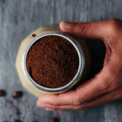 Mano che tiene la moka piena di caffè macinato, pronto per il rito del caffè. Chicchi sparsi sul tavolo di legno.