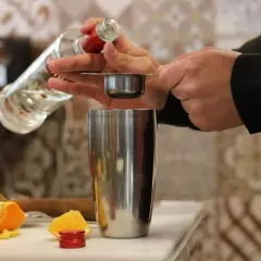 Preparazione di una ricetta cocktail: versando liquore da una bottiglia in un misurino sopra uno shaker, con spicchi d'arancia sullo sfondo.