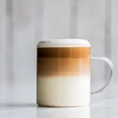 Un bicchiere di latte macchiato freddo con strati distinti di latte, caffè e schiuma, su uno sfondo sfocato.