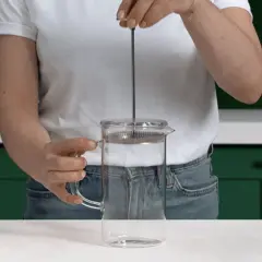 Mano che preme lo stantuffo di una french press di vetro, pronta per un caffè aromatico.