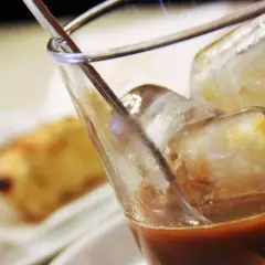 Un bicchiere di caffè con ghiaccio, perfetto per una pausa rinfrescante. Sullo sfondo, dolci sfocati invitano a un momento di piacere.