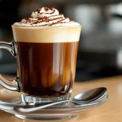 Un delizioso caffè marocchino con panna montata e cacao in polvere, servito in una tazza di vetro trasparente su un piattino con cucchiaino.