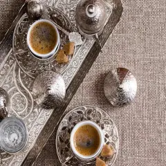 Un caffè in tazza grande, servito su un vassoio d'argento finemente decorato, con zollette di zucchero e bicchierino d'acqua.