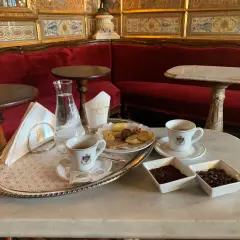 Un'elegante tazzina di caffè veneziano, biscotti e acqua su un vassoio d'argento, con divani rossi e decorazioni dorate sullo sfondo.
