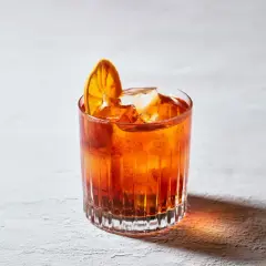 Un bicchiere di **bitter cocktail** arancione, con ghiaccio e una fetta d'arancia essiccata, su uno sfondo chiaro.