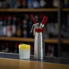 Sifone per panna montata accanto a un bicchiere con spuma drink giallo e bianco, su un bancone di bar con bottiglie sfocate sullo sfondo.