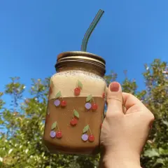 Un bicchiere di **iced coffee latte** decorato con ciliegie, tenuto in mano contro un cielo azzurro e alberi verdi.