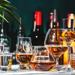Varietà di bicchieri con ambra liquida, che evocano le bevande tipiche italiane. Sullo sfondo, bottiglie scure e una foglia verde.