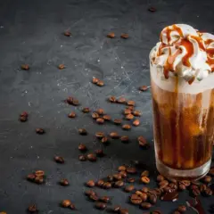 Un delizioso frappe al caffè con panna montata e sciroppo al caramello, circondato da chicchi di caffè tostati su sfondo scuro.