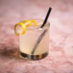 Un rinfrescante Vodka Sour IBA, guarnito con scorza di limone, su un tavolo di marmo rosa.