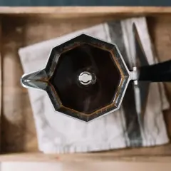 Moka nuova, appena usata, con caffè fumante, appoggiata su un panno a righe in una cassetta di legno.