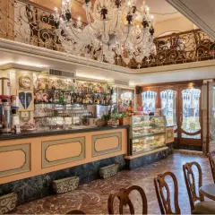 Interno di un caffè storico a Venezia, con bancone bar, vetrine di dolci, tavolini e un lampadario di cristallo.