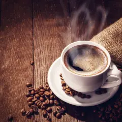 Tazza di caffè forte fumante su un piattino bianco, circondata da chicchi di caffè tostati su un tavolo di legno scuro.