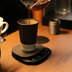 Versando caffè in una tazza scura, un'arte del coffee design. Bilancia digitale e filtro in primo piano.