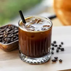 Un bicchiere di **cold brew latte** con ghiaccio e una cannuccia nera, accanto a una ciotola di chicchi di caffè tostati.