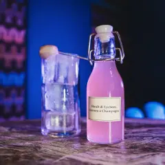 Bottiglia di shrub cocktail rosa con etichetta "Lychees, Zenzero e Champagne" accanto a un bicchiere pieno di ghiaccio.