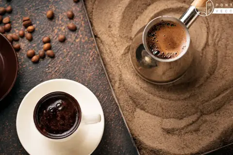 Un caffè arabo fumante in una tazzina bianca, accanto a un bricco di metallo su sabbia calda. Chicchi di caffè sparsi.