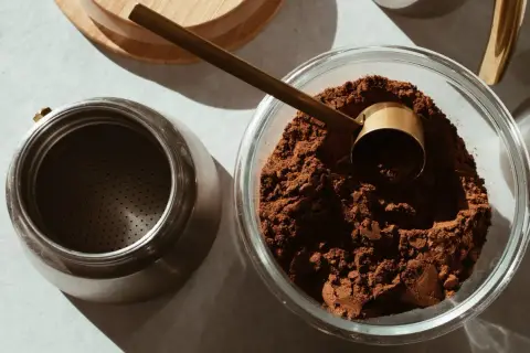 Caffè macinato in una ciotola di vetro, pronto per essere conservato. Un misurino dorato e una moka completano la scena, suggerendo come conservare il caffè al meglio.
