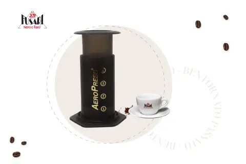 AeroPress per un caffè perfetto. Accanto, una tazzina bianca con logo e chicchi di caffè sparsi.