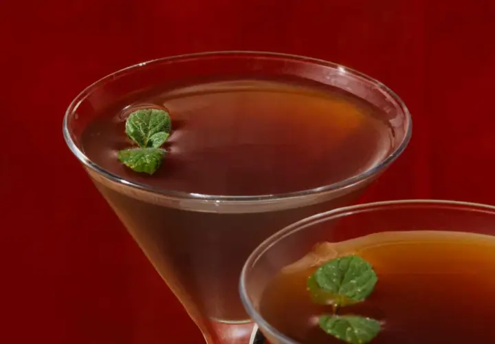 Due cocktail Stinger IBA, con liquore alla menta e brandy, guarniti con foglie di menta fresca, su sfondo rosso.