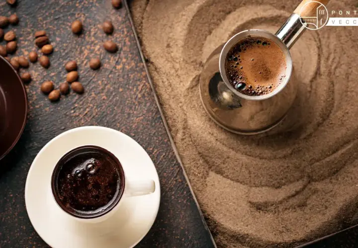 Un caffè arabo fumante in una tazzina bianca, accanto a un bricco di metallo su sabbia calda. Chicchi di caffè sparsi.