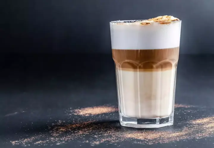 Un bicchiere di caffè macchiato caldo con strati di latte e caffè, sormontato da schiuma soffice e una spolverata di cacao.