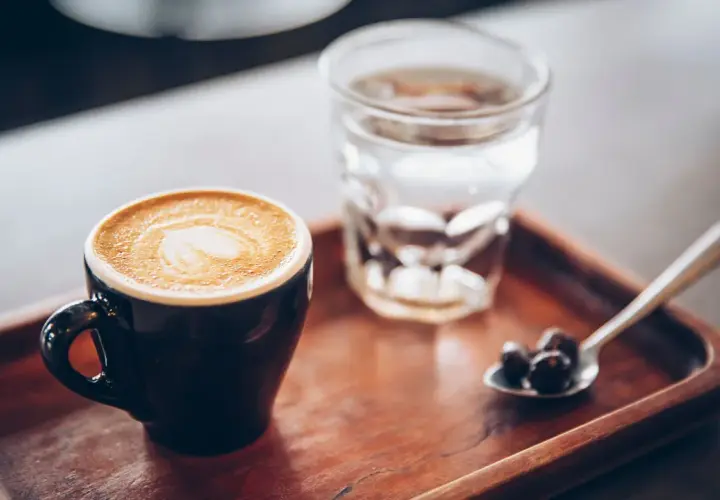 Tazza di caffè con latte art e bicchiere d'acqua, accompagnati da chicchi di caffè su un cucchiaino, su un vassoio di legno.