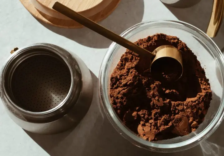 Caffè macinato in una ciotola di vetro, pronto per essere conservato. Un misurino dorato e una moka completano la scena, suggerendo come conservare il caffè al meglio.