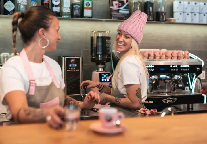 Due donne sorridono dietro il bancone di un bar, una con un cappello rosa, pronte a servire un caffè.