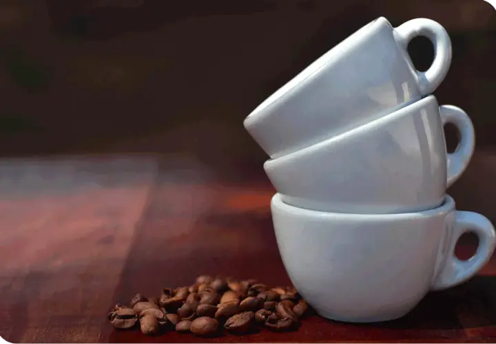 Tre tazzine bianche impilate, con chicchi di caffè tostati in primo piano. Immagina quanto contiene una tazzina di caffè.