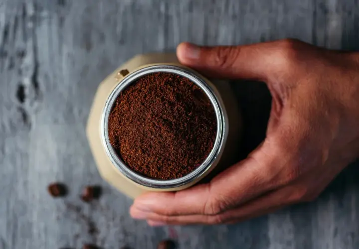 Mano che tiene la moka piena di caffè macinato, pronto per il rito del caffè. Chicchi sparsi sul tavolo di legno.