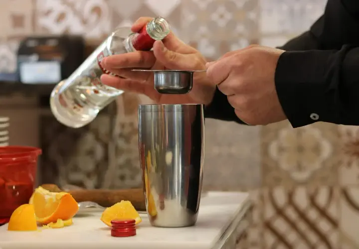 Preparazione di una ricetta cocktail: versando liquore da una bottiglia in un misurino sopra uno shaker, con spicchi d'arancia sullo sfondo.