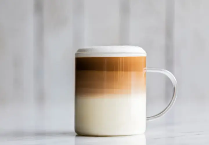 Un bicchiere di latte macchiato freddo con strati distinti di latte, caffè e schiuma, su uno sfondo sfocato.