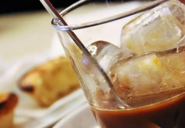 Un bicchiere di caffè con ghiaccio, perfetto per una pausa rinfrescante. Sullo sfondo, dolci sfocati invitano a un momento di piacere.