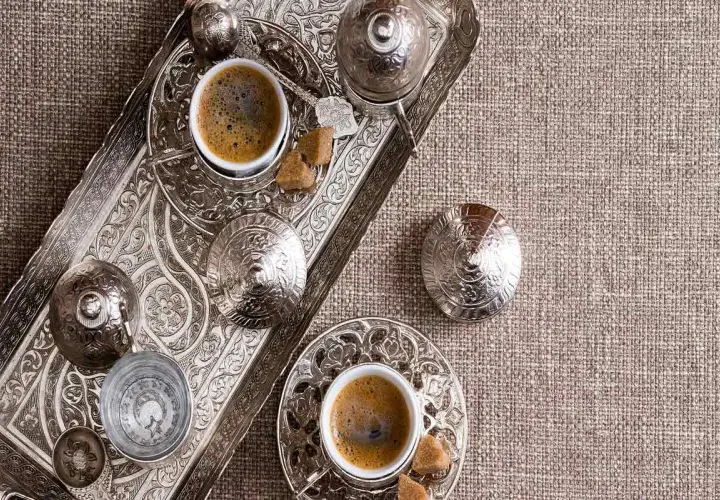 Un caffè in tazza grande, servito su un vassoio d'argento finemente decorato, con zollette di zucchero e bicchierino d'acqua.
