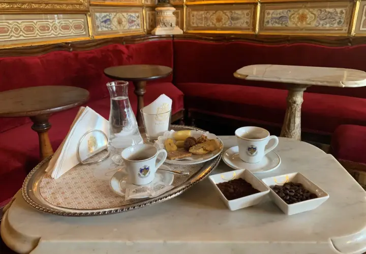 Un'elegante tazzina di caffè veneziano, biscotti e acqua su un vassoio d'argento, con divani rossi e decorazioni dorate sullo sfondo.