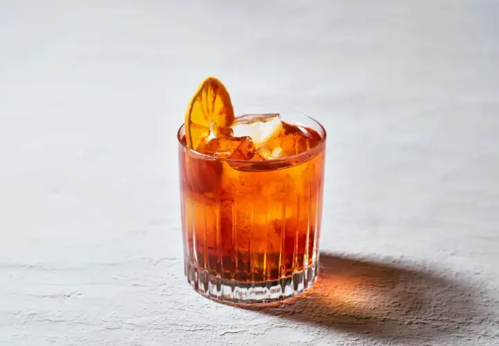 Un bicchiere di **bitter cocktail** arancione, con ghiaccio e una fetta d'arancia essiccata, su uno sfondo chiaro.
