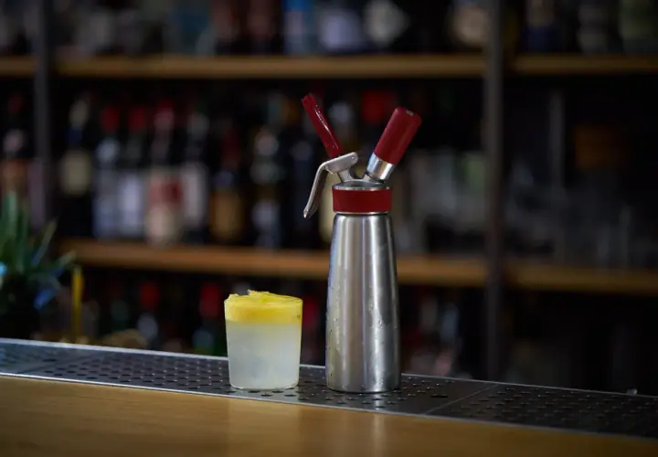 Sifone per panna montata accanto a un bicchiere con spuma drink giallo e bianco, su un bancone di bar con bottiglie sfocate sullo sfondo.