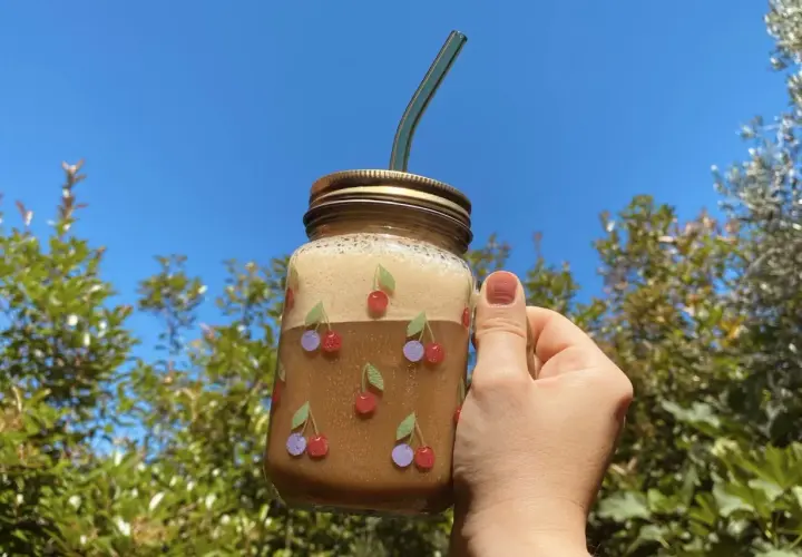 Un bicchiere di **iced coffee latte** decorato con ciliegie, tenuto in mano contro un cielo azzurro e alberi verdi.
