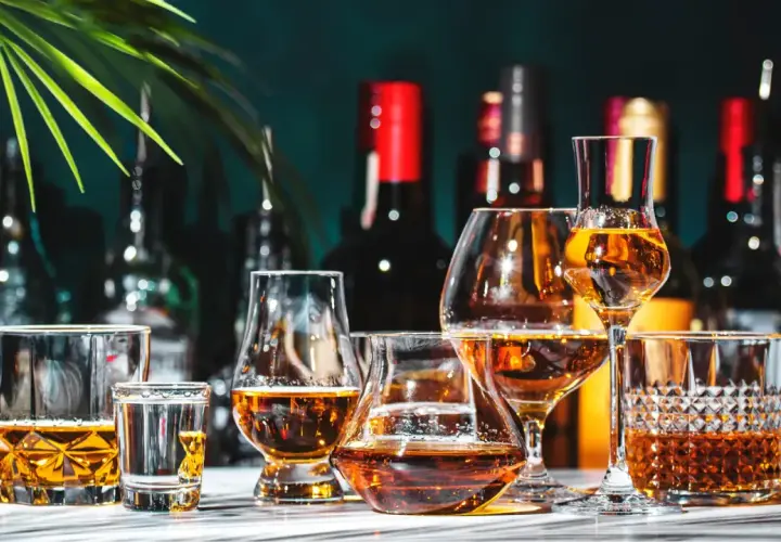 Varietà di bicchieri con ambra liquida, che evocano le bevande tipiche italiane. Sullo sfondo, bottiglie scure e una foglia verde.