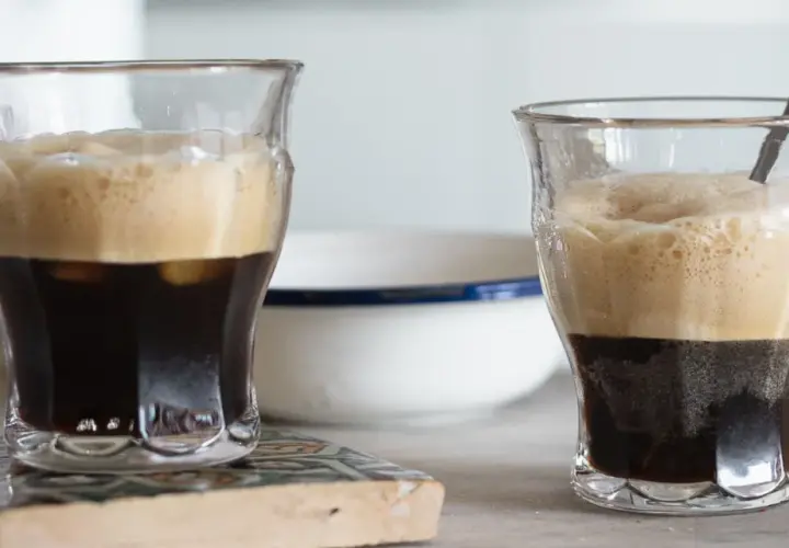 Due bicchieri di caffè shakerato cremoso, con la schiuma dorata che sovrasta il caffè scuro. Uno ha un cucchiaino.