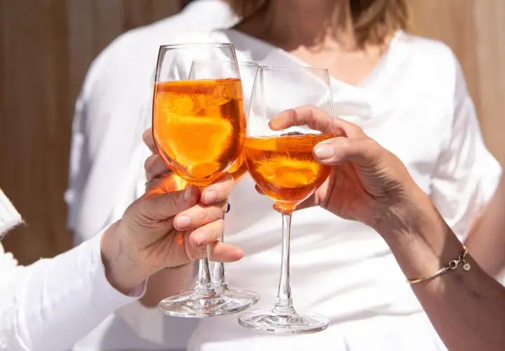 Tre mani brindano con bicchieri di Aperol, un drink rinfrescante e senza glutine, sotto il sole.