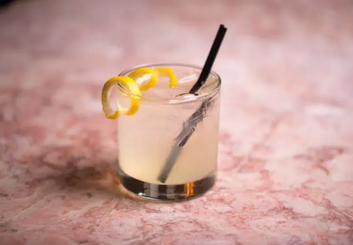 Un rinfrescante Vodka Sour IBA, guarnito con scorza di limone, su un tavolo di marmo rosa.