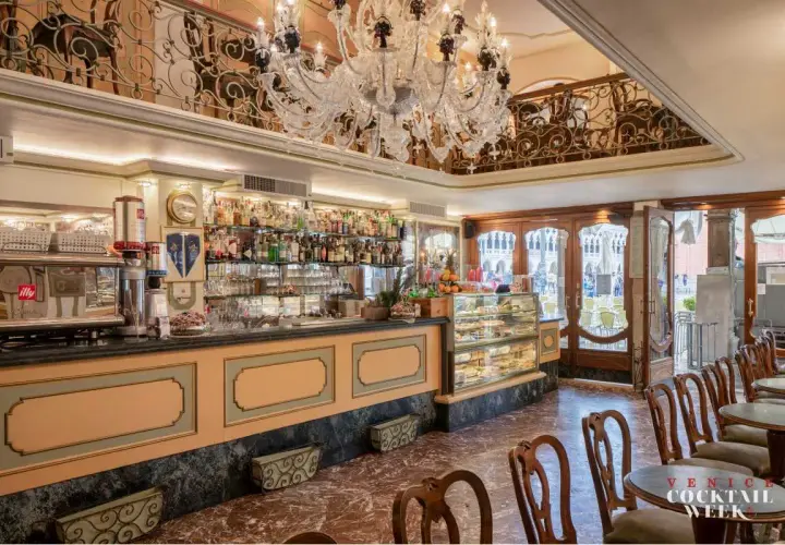 Interno di un caffè storico a Venezia, con bancone bar, vetrine di dolci, tavolini e un lampadario di cristallo.