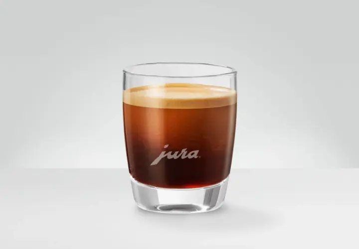 Un bicchiere di vetro con un doppio macchiato, la schiuma dorata corona il caffè scuro. Il logo Jura è inciso sul vetro.